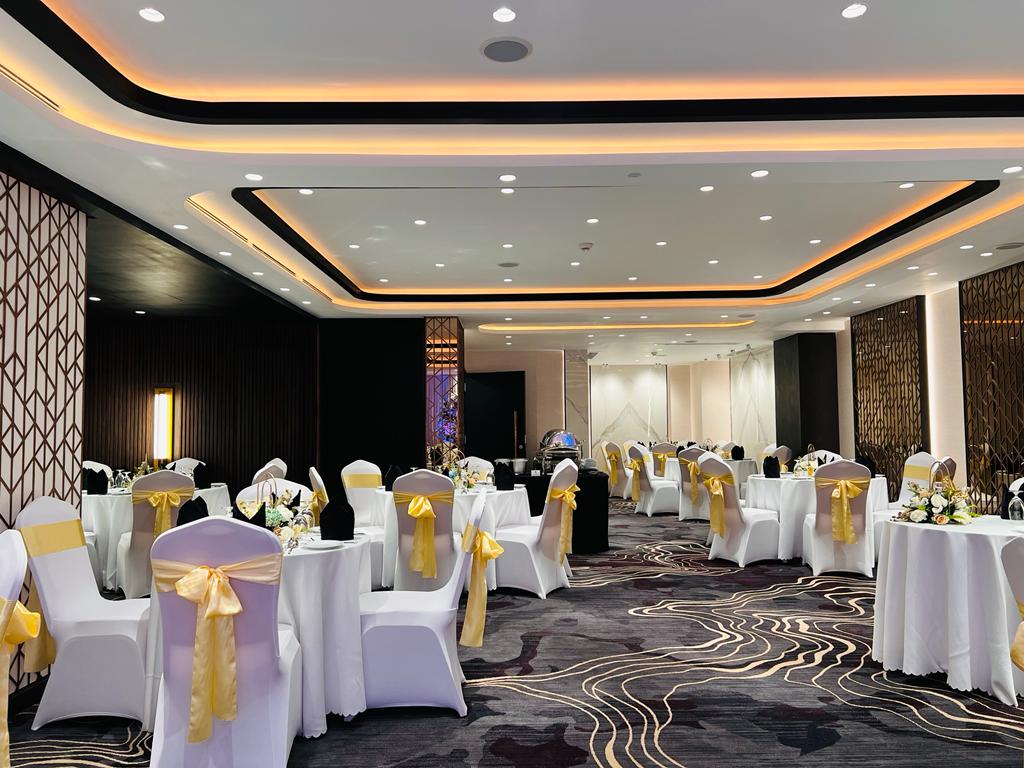 Elegant banquet hall with white chair covers, yellow sashes, decorated tables, and modern lighting.